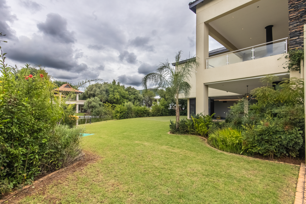 A spacious rear garden featuring a manicured lawn, established landscaping, and a covered patio area with a built-in fireplace overlooking the estate grounds.