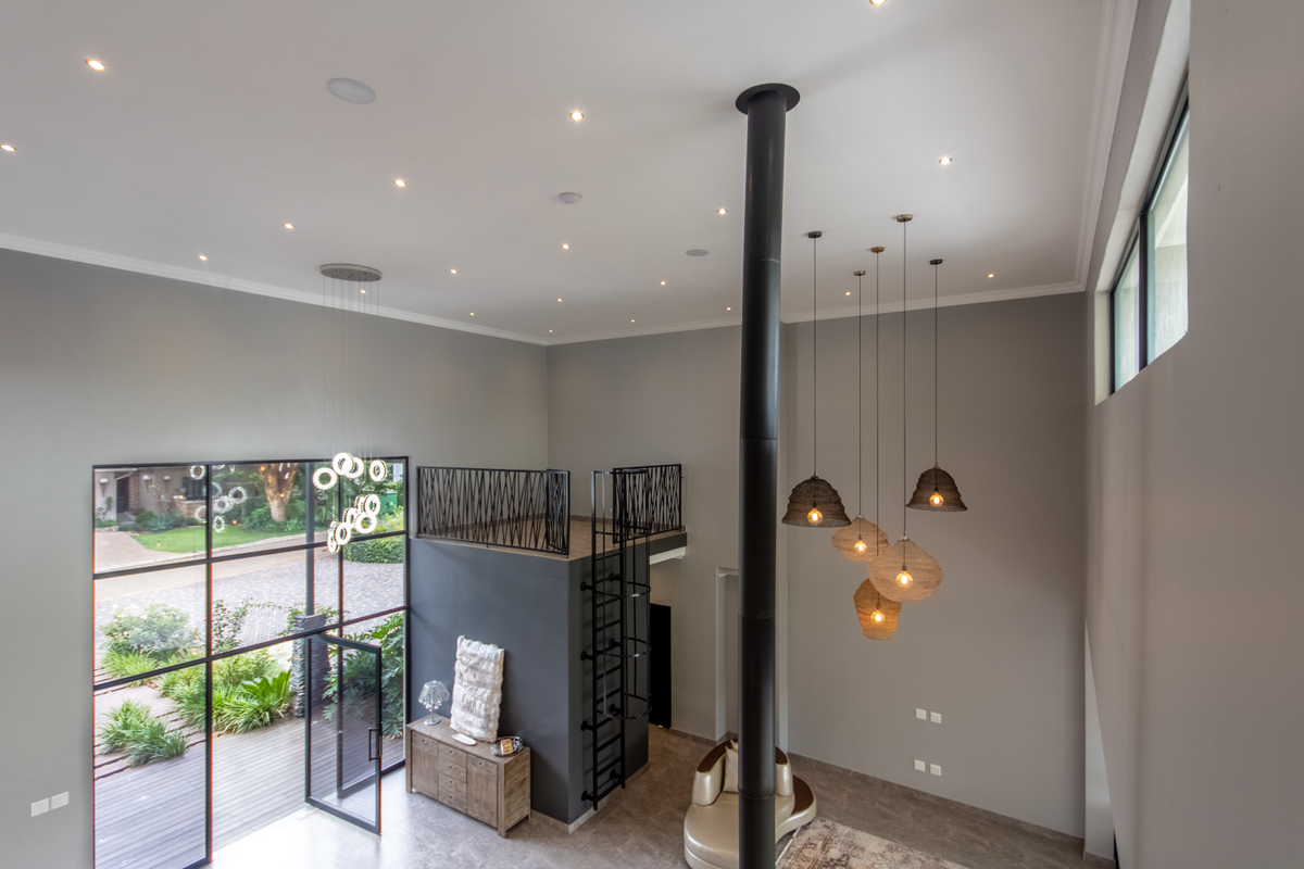 Spacious double-volume living area featuring a mezzanine level, industrial-style metal staircase, floor-to-ceiling glass windows, and a central fireplace flue.