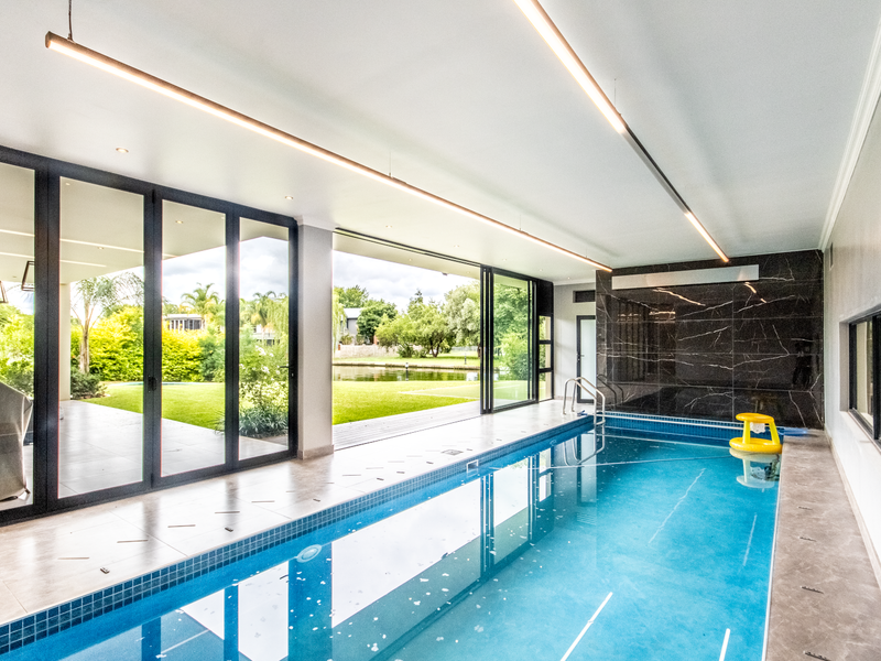 Indoor lap pool area featuring large sliding glass doors opening to a landscaped garden and water view, with modern linear lighting and tiled surrounds.