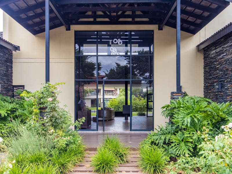 A striking modern entrance featuring a high-pitched roof, expansive glass facade, and natural stone cladding, framed by lush, well-maintained garden landscaping.