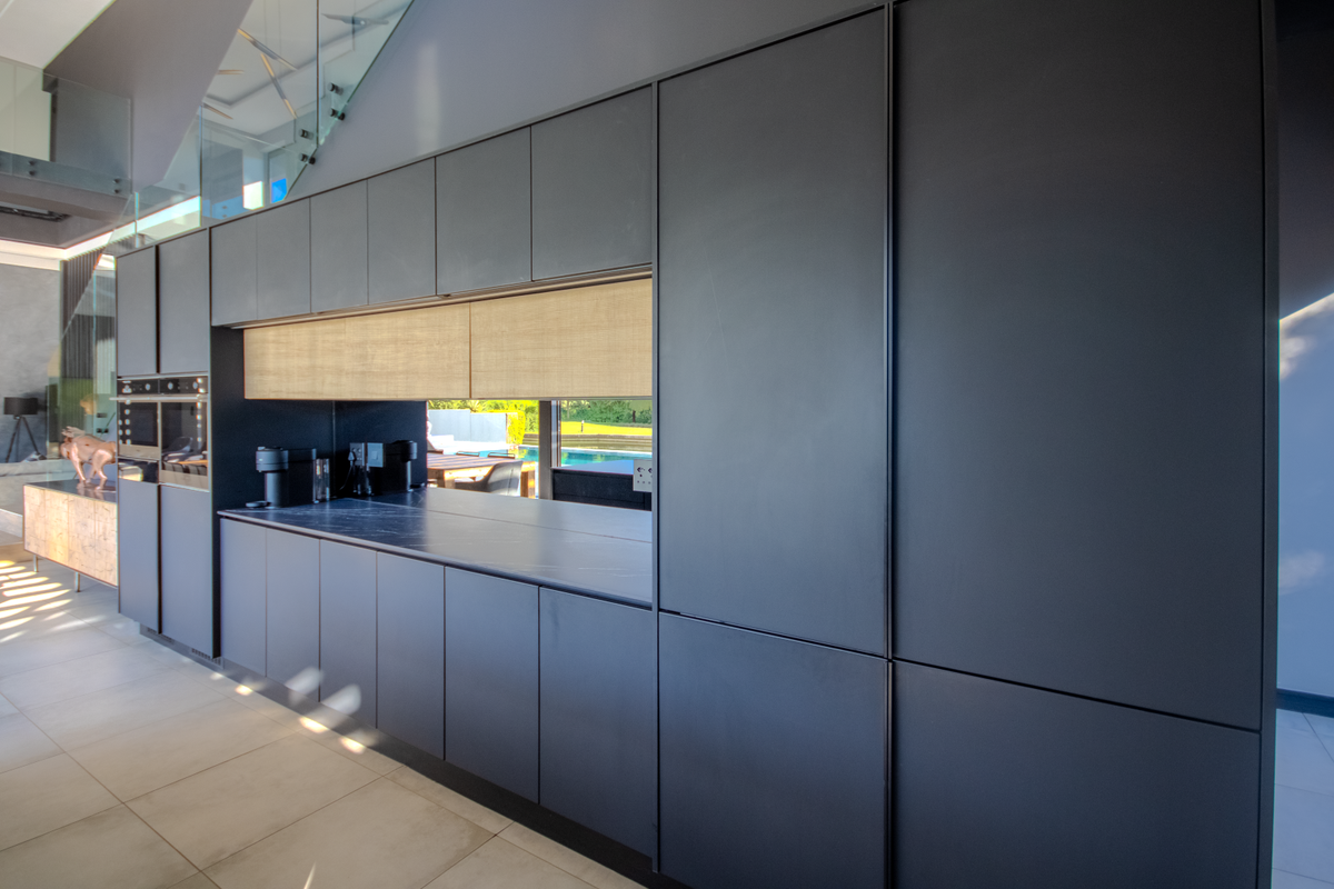 Modern kitchen featuring sleek matte dark cabinetry, integrated appliances, and a stone countertop with a pass-through window overlooking the outdoor pool area.