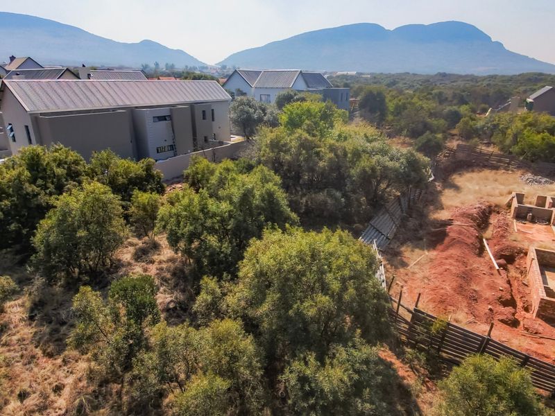Aerial view of a residential vacant land plot featuring established trees, perimeter fencing, and an active foundation construction site with mountain views.