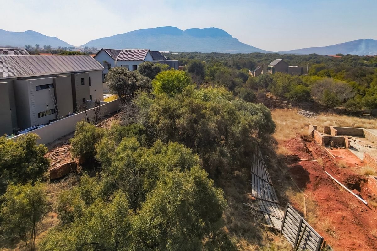 Aerial view of a residential plot featuring native vegetation, a brick foundation under construction, and surrounding neighborhood development with mountain views.
