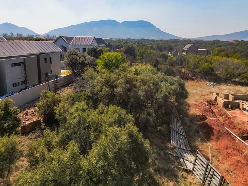 Aerial view of a residential plot featuring native vegetation, a brick foundation under construction, and surrounding neighborhood development with mountain views.