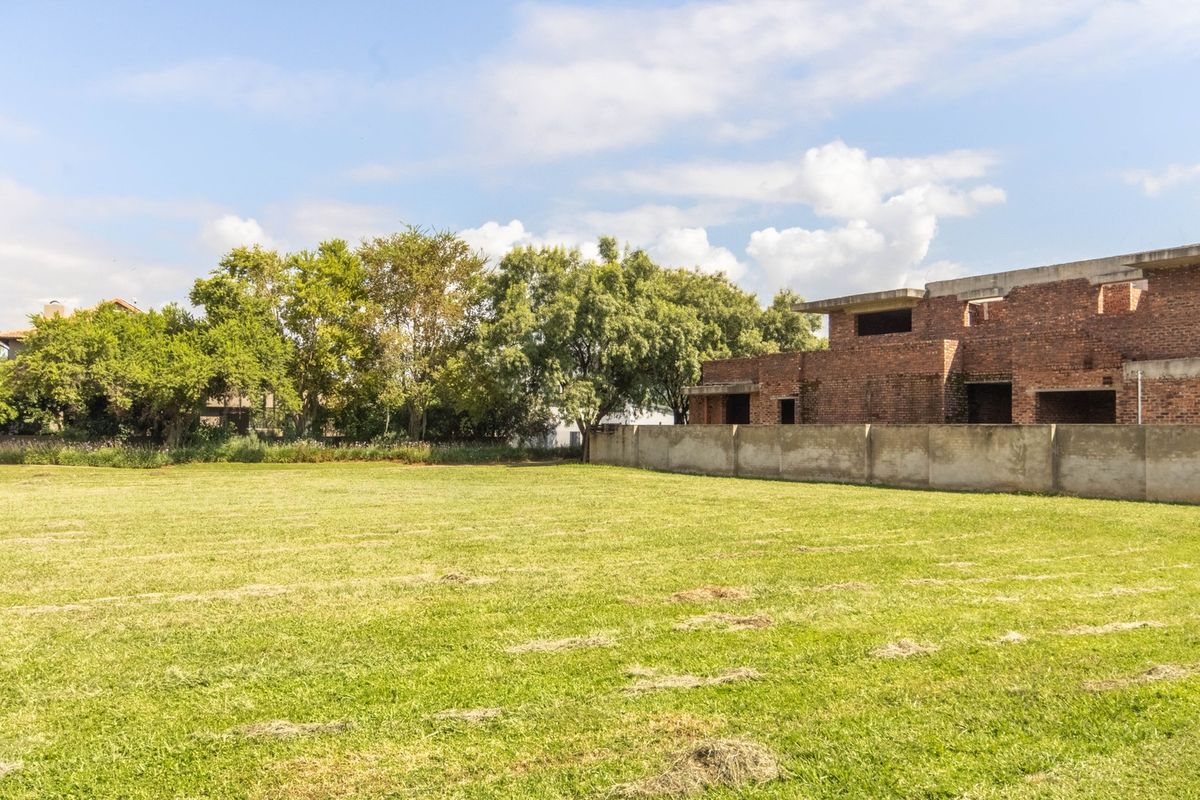 A spacious vacant residential lot featuring a large grassed area, boundary wall, and an adjacent unfinished brick building structure.