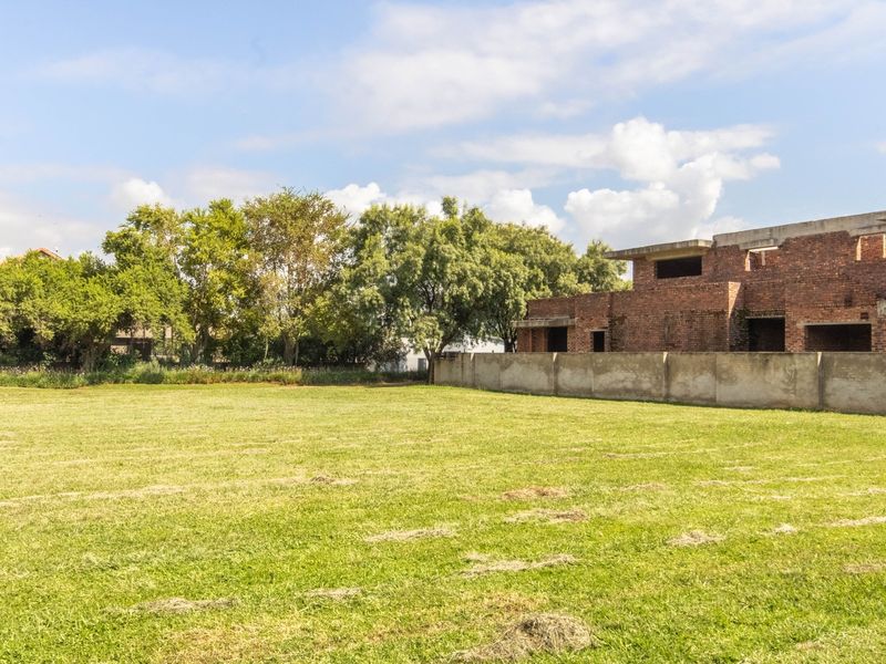 A spacious vacant residential lot featuring a large grassed area, boundary wall, and an adjacent unfinished brick building structure.