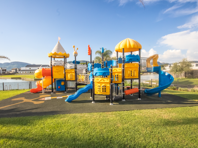 A well-maintained outdoor playground facility featuring colorful climbing structures, slides, and safety surfacing, situated within a residential estate near the water.