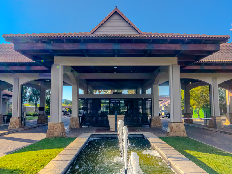 A well-maintained residential estate entrance featuring a grand covered portico, stone-clad pillars, and a central water fountain.