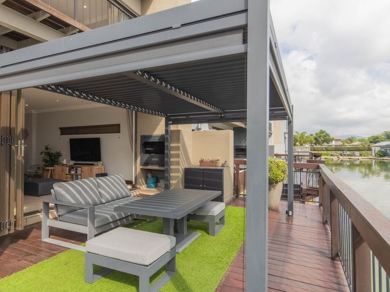 A modern covered patio featuring a louvered pergola, built-in braai, and timber decking, offering direct access to the living area and waterfront views.