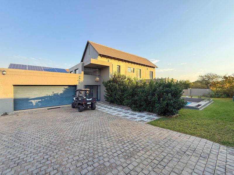 Front elevation of a modern house featuring a paved driveway, integrated garage, solar panel installation, and a glimpse of the side garden and pool area.