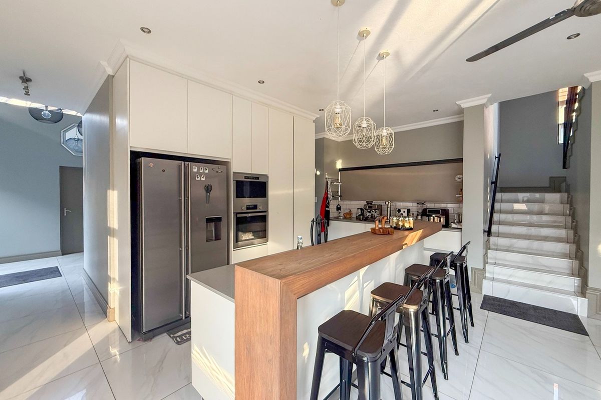 Modern kitchen featuring sleek white cabinetry, a wooden breakfast bar, integrated appliances, and polished tiled flooring, adjacent to a staircase.