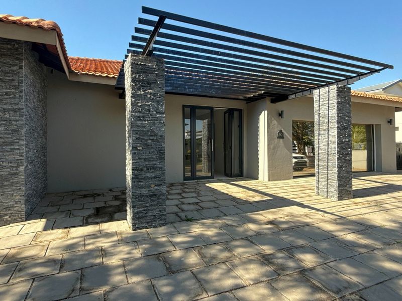 Front entrance featuring a modern metal pergola, stone-clad pillars, and a paved patio area leading to bi-fold glass doors.