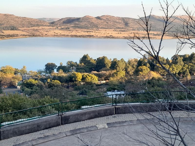 Elevated view overlooking a lake and surrounding landscape, featuring a paved area with a metal safety railing.
