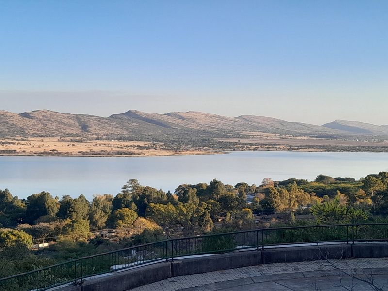A scenic elevated view overlooking a large lake, surrounding trees, and distant mountain ranges from a paved terrace with a metal safety railing.