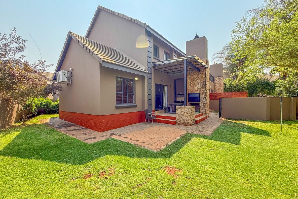 Rear view of the townhouse featuring a paved patio area with a built-in braai, stone cladding accents, and a well-maintained garden.