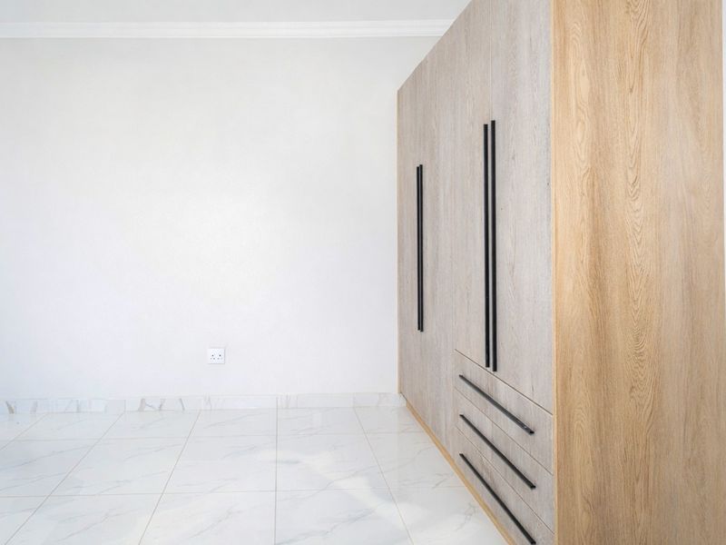 A bright, empty bedroom featuring modern light wood built-in cupboards with black handles and polished marble-look floor tiles.