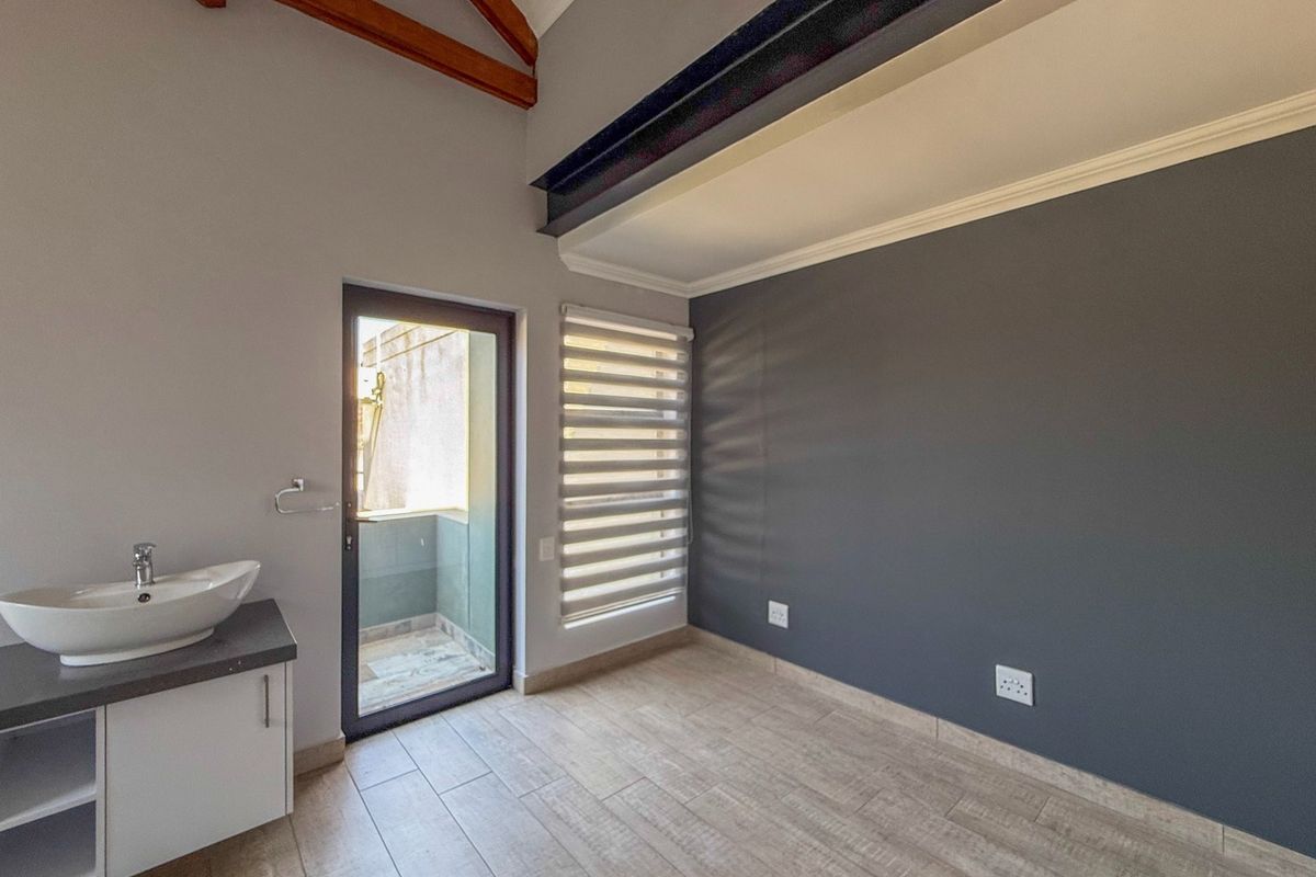 A modern bedroom featuring exposed timber rafters, a steel beam, tiled flooring, and an en-suite vanity basin with direct access to a small balcony.
