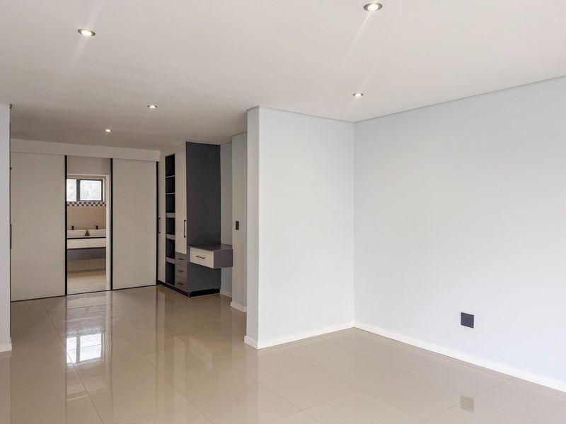 Spacious bedroom featuring neutral tiled flooring, recessed downlights, and built-in cabinetry with an integrated vanity, leading into an en-suite bathroom area.