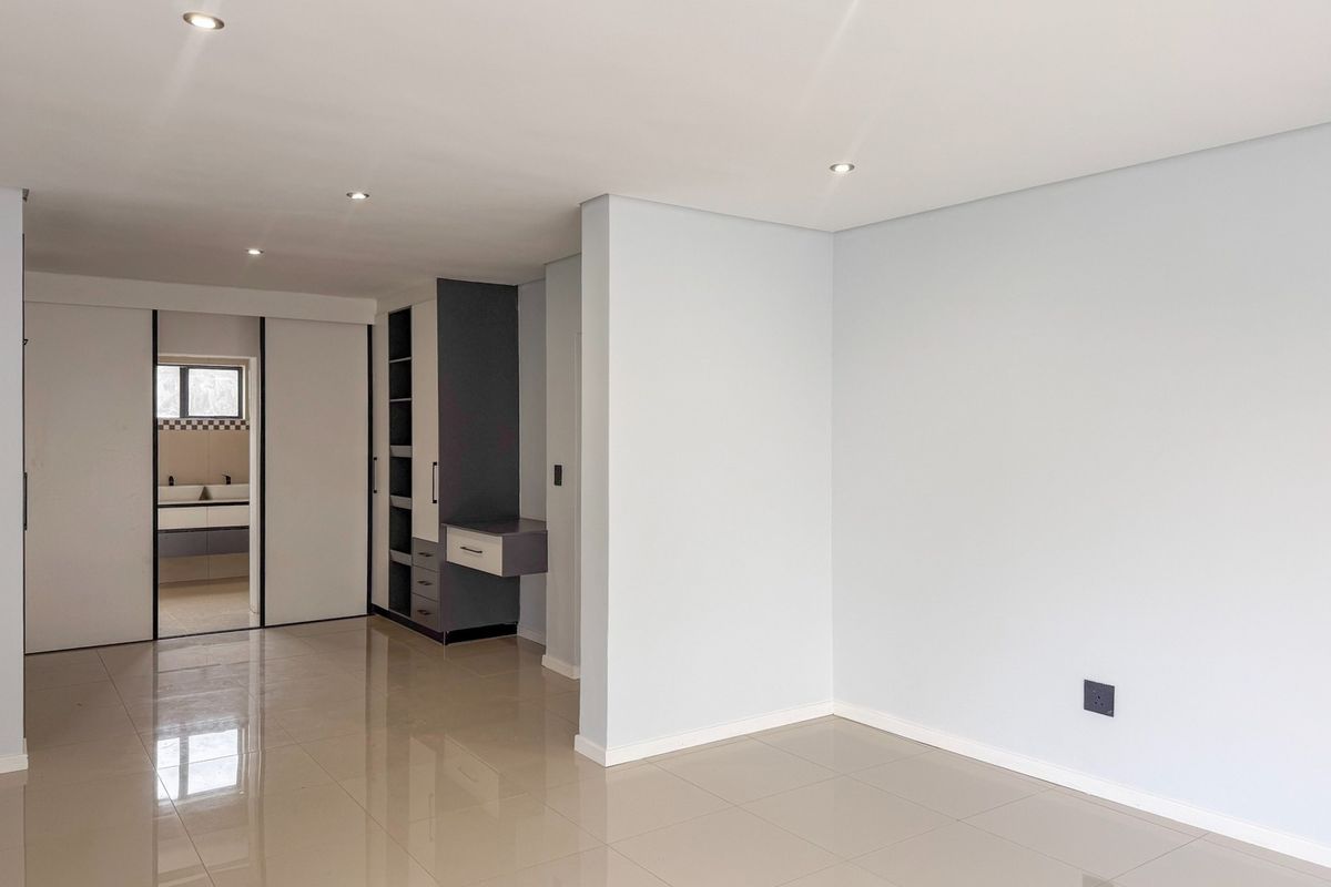 Spacious bedroom featuring neutral tiled flooring, recessed downlights, and built-in cabinetry with an integrated vanity, leading into an en-suite bathroom area.