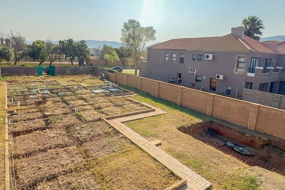 Aerial view of a residential plot under construction, featuring perimeter walls, brick pathways, and an excavated area prepared for foundation work.