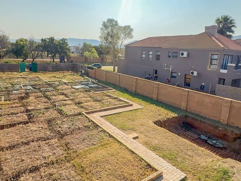 Aerial view of a residential plot under construction, featuring perimeter walls, brick pathways, and an excavated area prepared for foundation work.