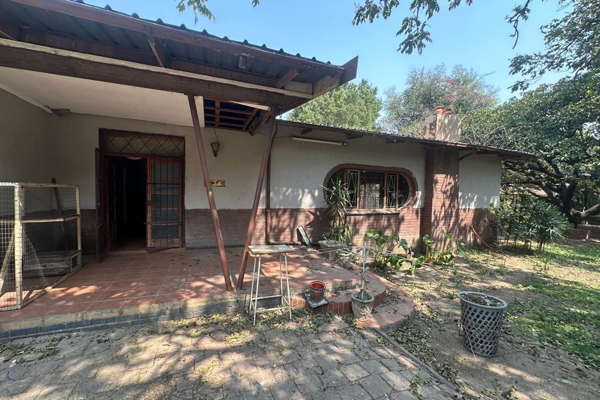 Rear exterior view of the property featuring a covered patio area, brick facade, and established garden surroundings requiring maintenance.