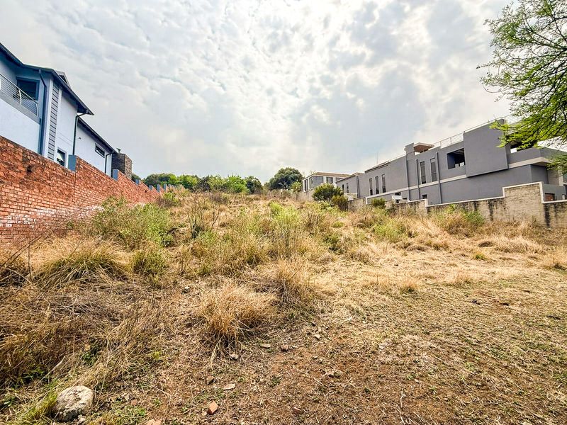 A vacant residential plot featuring a sloped landscape, enclosed by brick boundary walls and situated between existing modern residential developments.