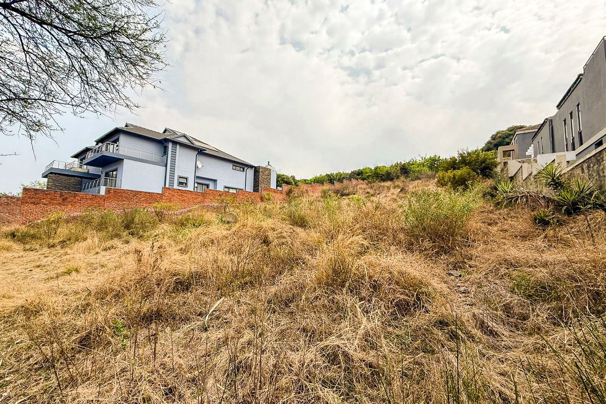 A view of a sloped, undeveloped residential plot featuring dry vegetation and a brick boundary wall, adjacent to existing multi-story residential properties.