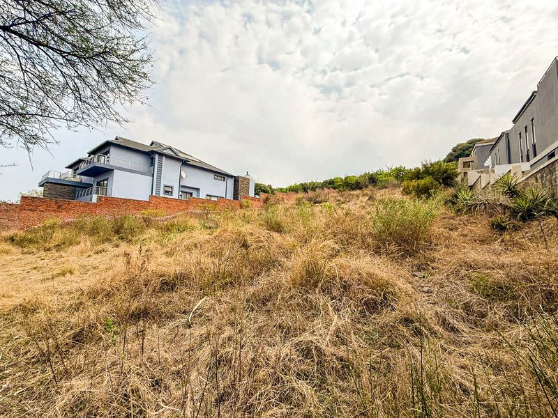 A view of a sloped, undeveloped residential plot featuring dry vegetation and a brick boundary wall, adjacent to existing multi-story residential properties.