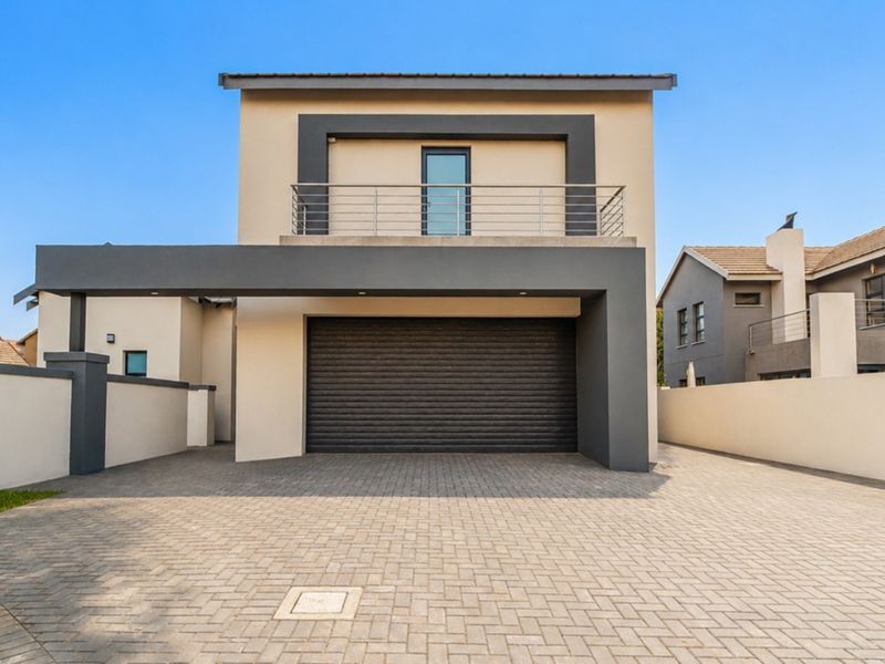 Modern double-story residential facade featuring a paved driveway, automated garage door, and an upper-level balcony with contemporary metal railings.