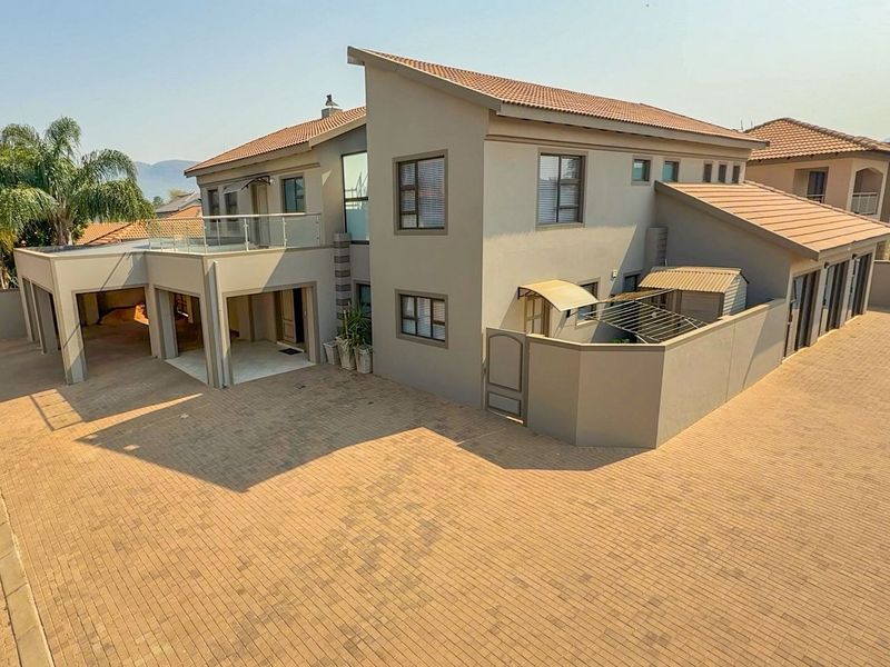 A modern double-story residential property featuring a paved driveway, covered patio, and an upper-level balcony with glass balustrades.