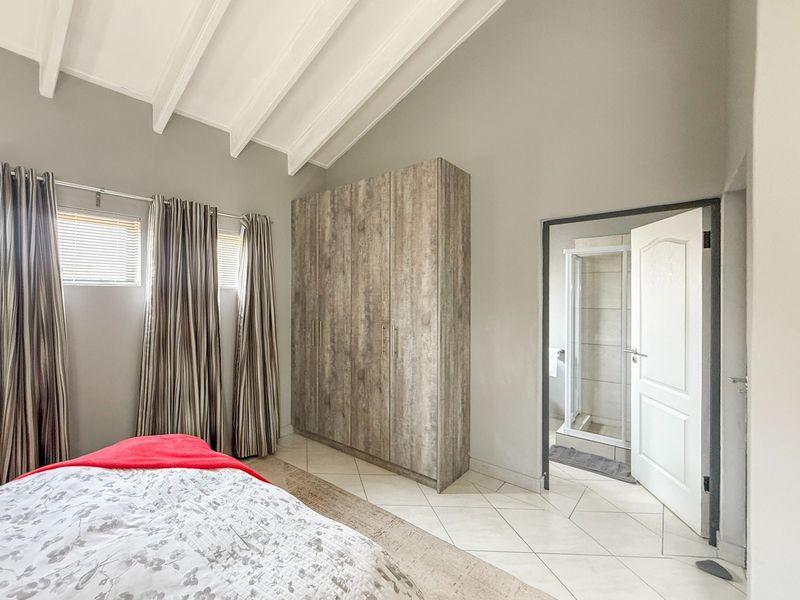 A well-maintained bedroom featuring vaulted ceilings with exposed beams, built-in wooden wardrobes, tiled flooring, and direct access to an en-suite bathroom.