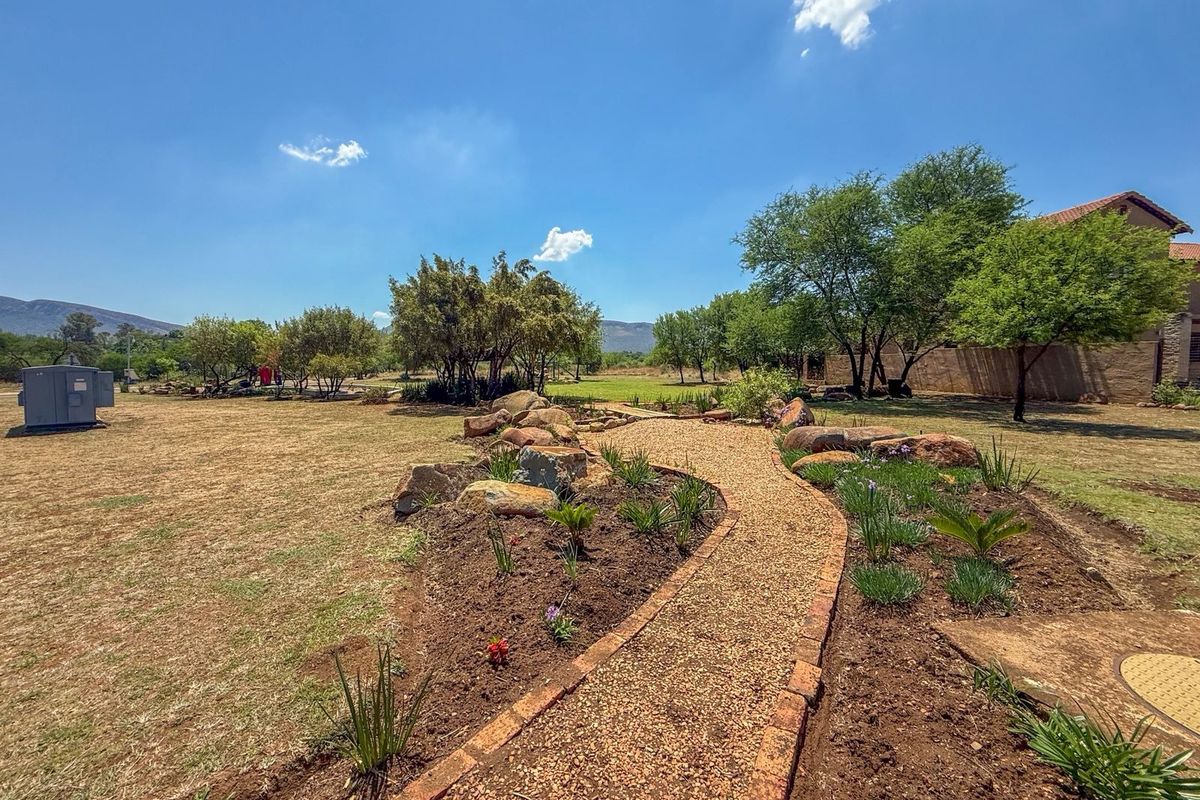 A well-maintained garden area featuring a gravel pathway with brick edging, decorative boulder rockery, and established trees on a spacious residential plot.