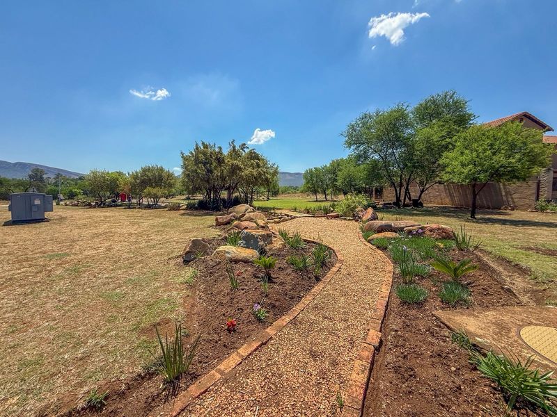 A well-maintained garden area featuring a gravel pathway with brick edging, decorative boulder rockery, and established trees on a spacious residential plot.