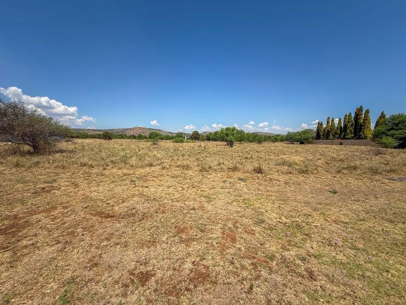 A wide-angle view of a vacant residential plot featuring natural dry grass, scattered trees, and distant mountain views.