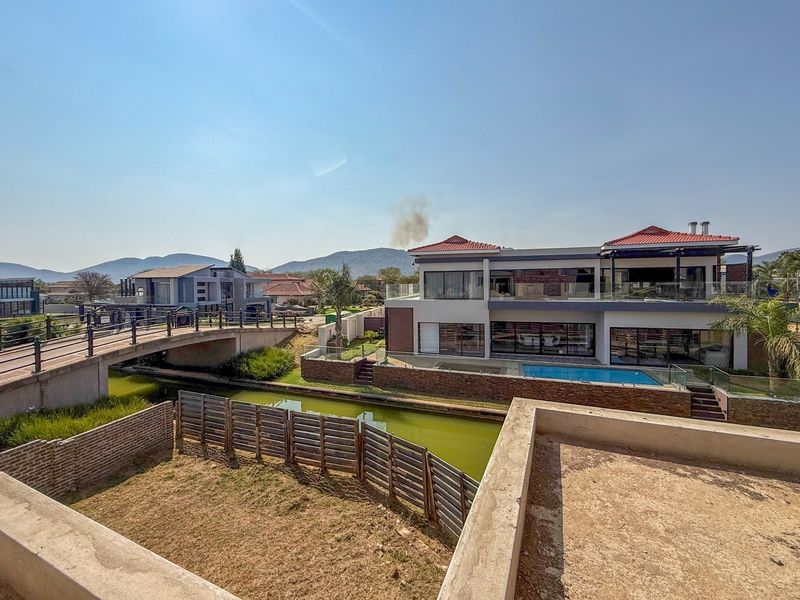 Rear view of a modern multi-story house featuring a swimming pool, stone cladding, and glass balustrades, overlooking a canal and bridge.