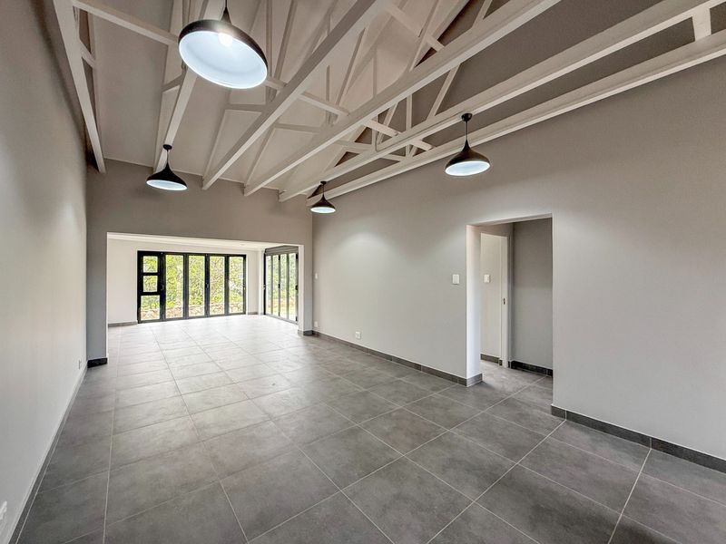 Spacious open-plan living area featuring high ceilings with exposed white trusses, modern grey floor tiles, and large glass doors leading to the outdoors.