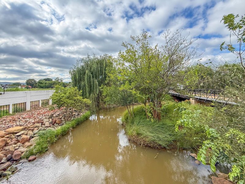 A scenic view of a landscaped riverbank featuring a stone retaining wall, mature trees, and a pedestrian bridge within the retirement village grounds.