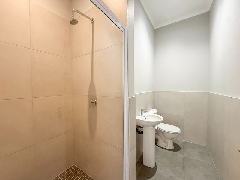 A clean, modern bathroom featuring a glass-enclosed shower, pedestal basin, and toilet with neutral wall and floor tiling.
