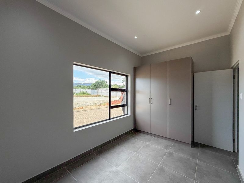 A modern, empty bedroom featuring neutral grey walls, large format floor tiles, recessed lighting, and a built-in wardrobe with a view of the exterior.