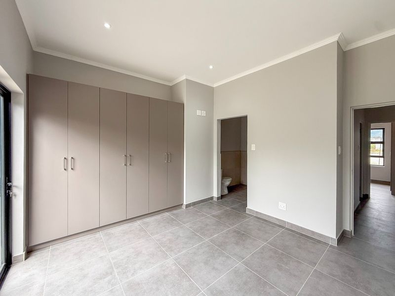 A bright, modern bedroom featuring neutral grey floor tiles, ample built-in cupboard storage, and direct access to an en-suite bathroom.