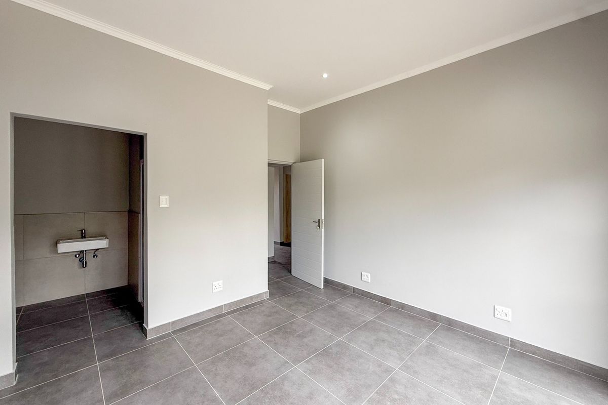 A clean, modern, and empty bedroom featuring neutral grey tiled flooring, recessed downlighting, and an adjoining small wash area with a wall-mounted basin.