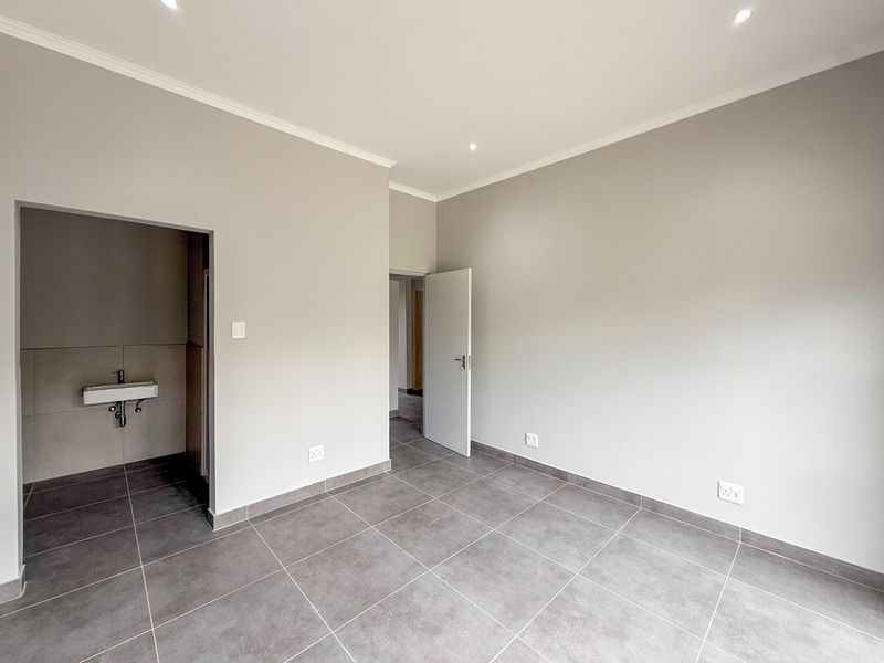 A clean, modern, and empty bedroom featuring neutral grey tiled flooring, recessed downlighting, and an adjoining small wash area with a wall-mounted basin.