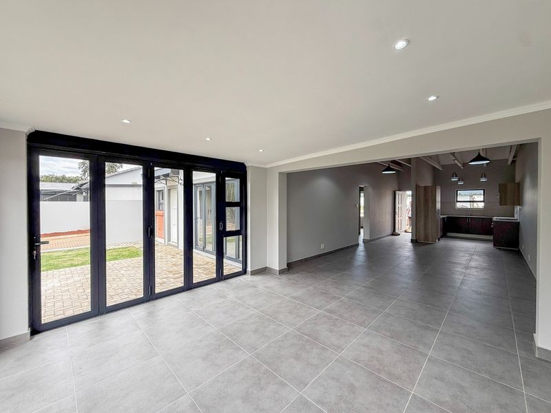 Spacious open-plan living area featuring large black aluminium stacker doors leading to a paved patio, with an adjoining kitchen and modern tiled flooring.