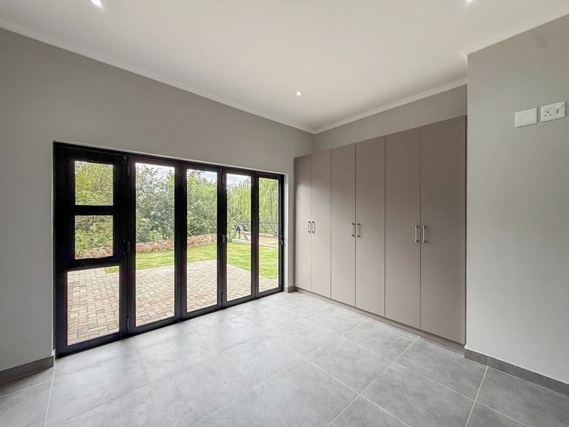 A bright, modern bedroom featuring neutral tiled flooring, ample built-in cupboard space, and large aluminium stacker doors opening onto a private garden patio.