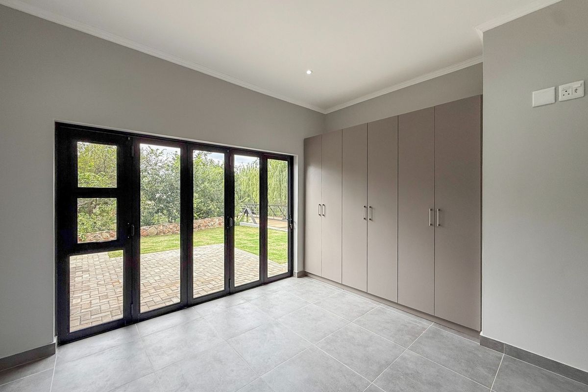 A bright, modern bedroom featuring neutral tiled flooring, ample built-in cupboard space, and large aluminium stacker doors opening onto a private garden patio.