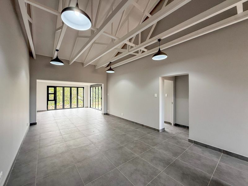 Spacious open-plan living area featuring high ceilings with exposed white trusses, modern grey floor tiles, and large black aluminium stacker doors leading outdoors.