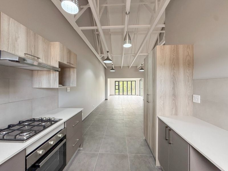 Modern open-plan kitchen and living area featuring light wood cabinetry, gas hob, exposed white roof trusses, and large glass doors leading to the exterior.