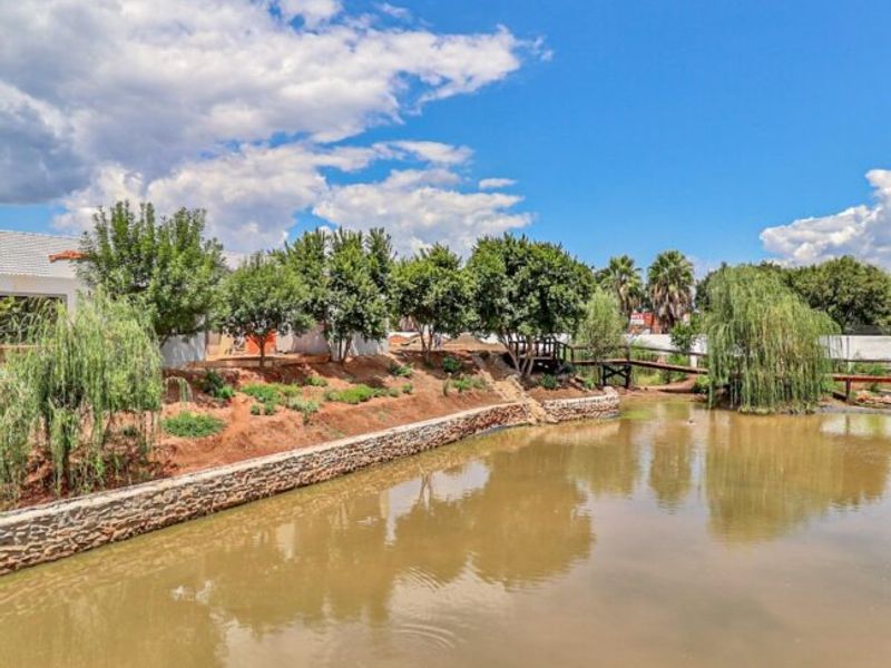 A scenic view of a landscaped garden featuring a stone retaining wall, a small pond, and a wooden footbridge within the retirement village grounds.