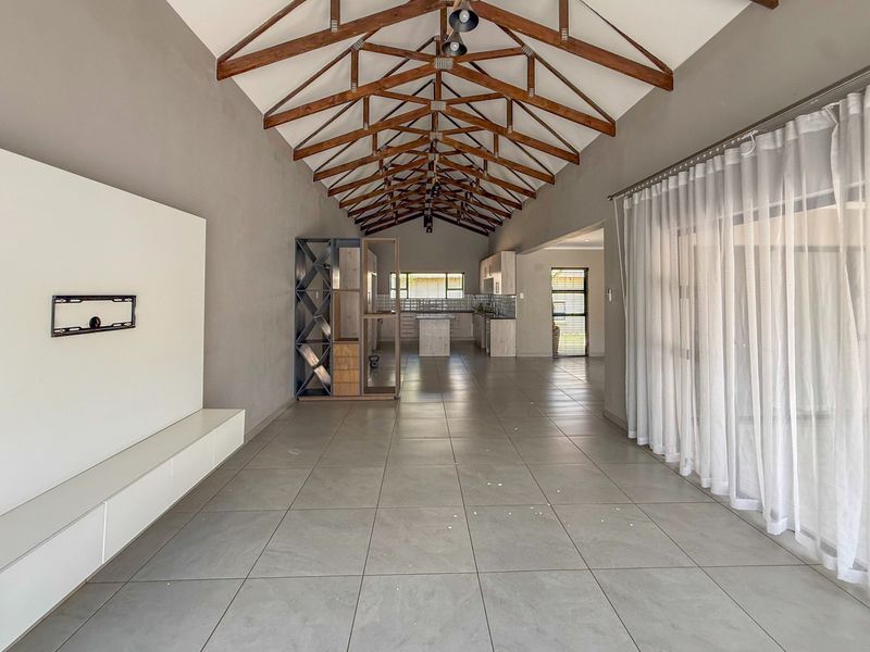 Spacious open-plan living and kitchen area featuring high vaulted ceilings with exposed timber trusses, large format floor tiles, and modern built-in cabinetry.
